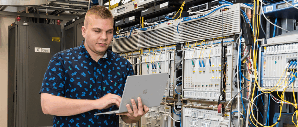 A student holds a laptop and runs a diagnostic on the network servers behind him.