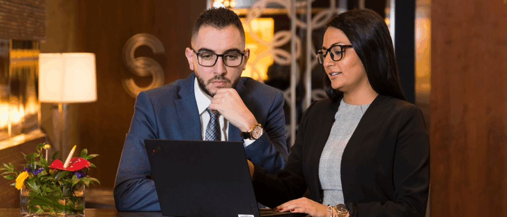 Two students dressed in business suits look at a screen at the reception desk of a hotel.