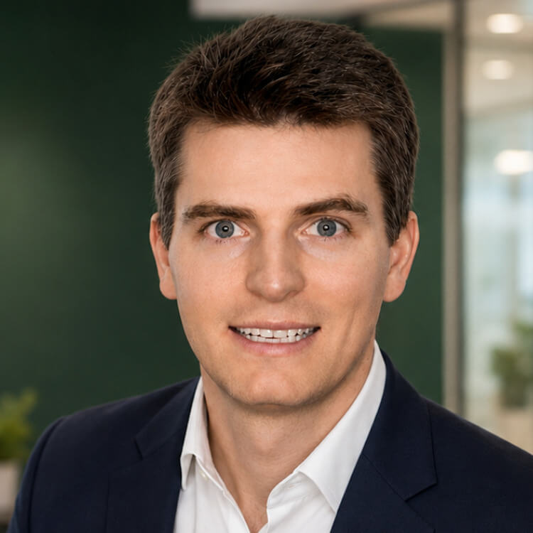Dr. J. (Ian) Zinck - A person with short brown hair wearing a dark suit jacket and white collared shirt stands in a bright office setting with green walls, plants, and glass partitions softly blurred in the background.