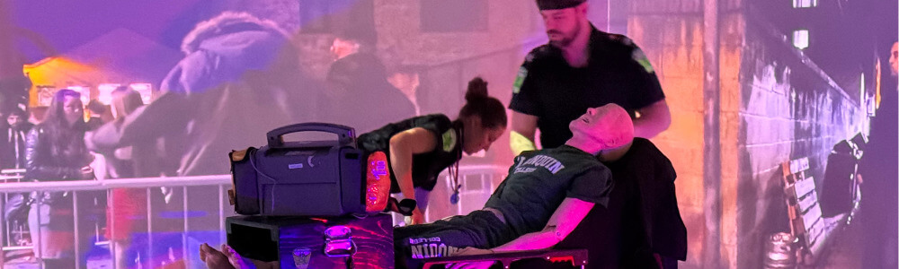 Two students training to be emergency responders assist a person lying on a medical stretcher, with medical equipment beside them and a projected urban scene in purple and pink tones in the background.