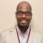 A person stands in front of a plain beige wall, smiling at the camera. They wear glasses, a light-colored blazer over a light blue collared shirt, and a red lanyard. The photo is a simple, professional portrait.