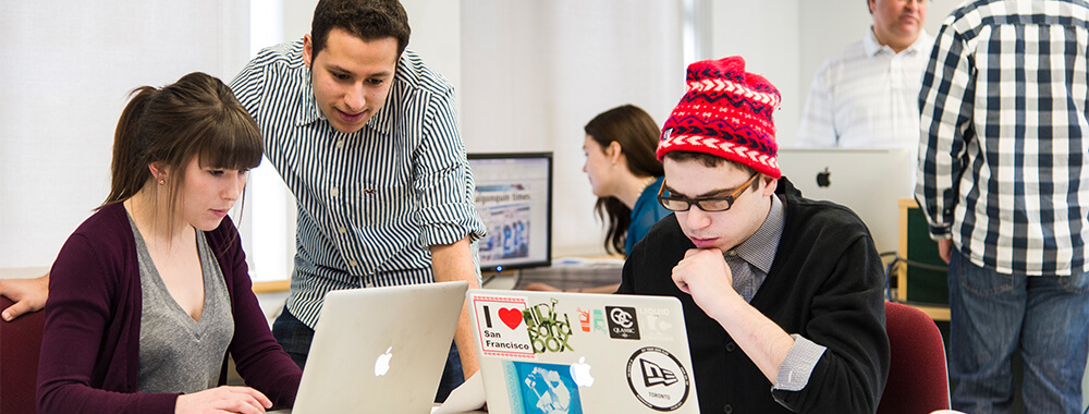 College classroom with three students collaborating at a table—two seated and working on laptops, while a third stands and looks at one of the screens. In the background, another student is focused on a desktop monitor, and a different student is speaking with the professor.