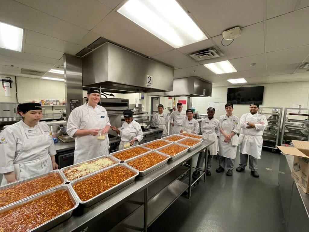 Many students in a kitchen posing for a photo behind a large amount of food that they are preparing.