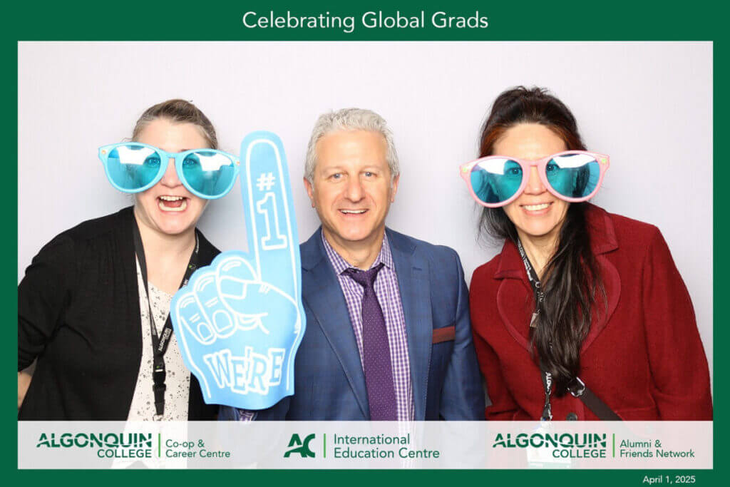 Three people - two women and one man in the middle - smile while wearing oversized sunglasses and a foam "we're number 1" finger 