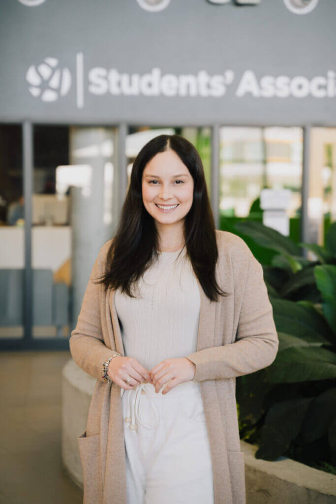 A woman with long dark hair stands in front of a sign that says Students' Association.