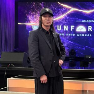 A man wearing black stands in front of a purple digital screen.