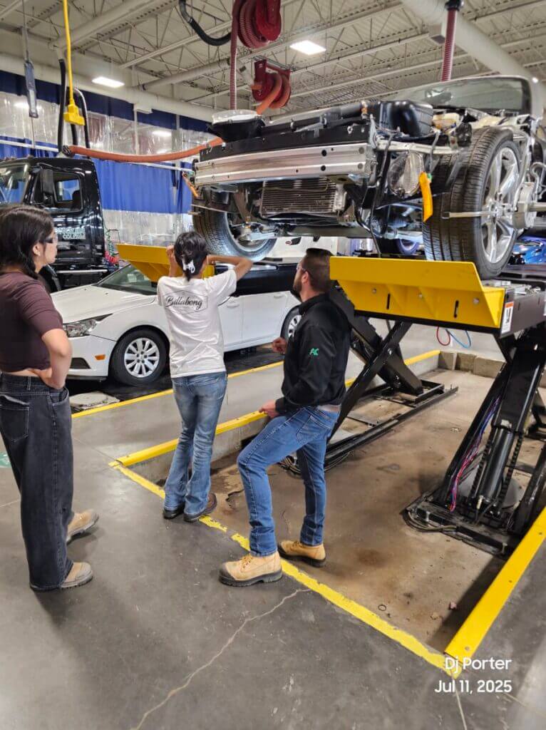 Two students and an instructor look at a car on an auto lift.