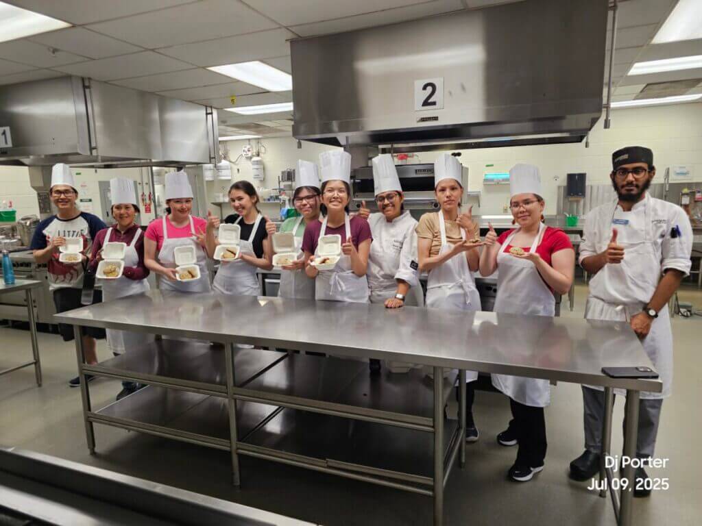 Students wear aprons and chef hands in a professional kitchen, holding cookies, salsa and guacamole. 