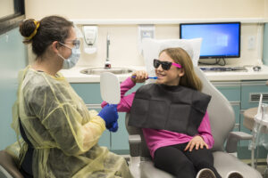 A woman showing a child how to brush their teeth in a dental office using a mirror.