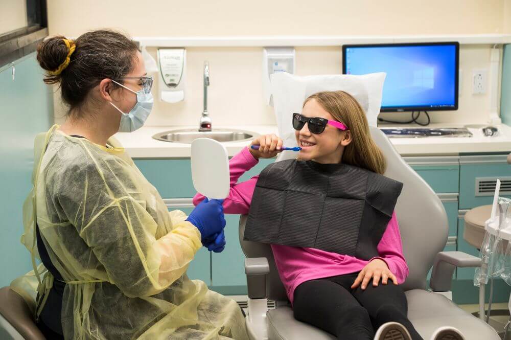 A woman showing a child seated in a dental chair in a clinic how to brush their teeth using a mirror.