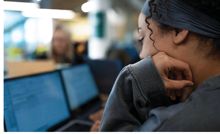 A woman is sitting at a desk with a laptop in front of her. She is wearing a gray sweatshirt and has her hand on her chin. AI generated content