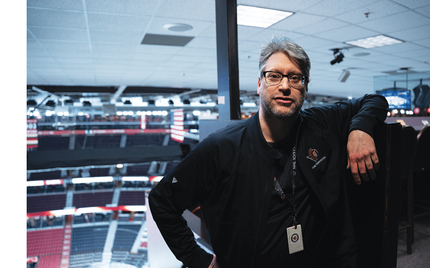 A man wearing glasses and a black jacket is standing in a stadium. AI generated content