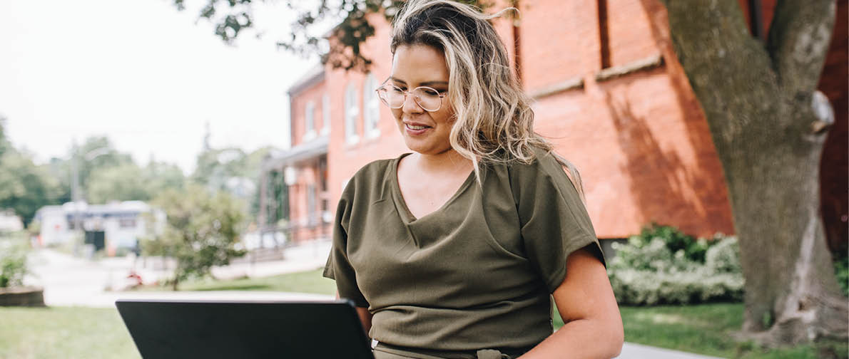 A woman is sitting outside on a sidewalk, using a laptop computer. AI generated content