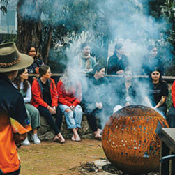 A group of people are gathered around a fire pit, with some sitting on a bench and others standing. A man in a hat is standing near the fire pit, and a woman in a red shirt is sitting on the bench. The group appears to be enjoying a social event together. AI generated content