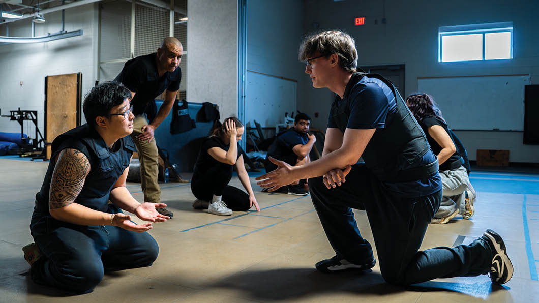 A group of people are in a room, with one man kneeling down and another man standing over him. The standing man is holding a remote control. AI generated content