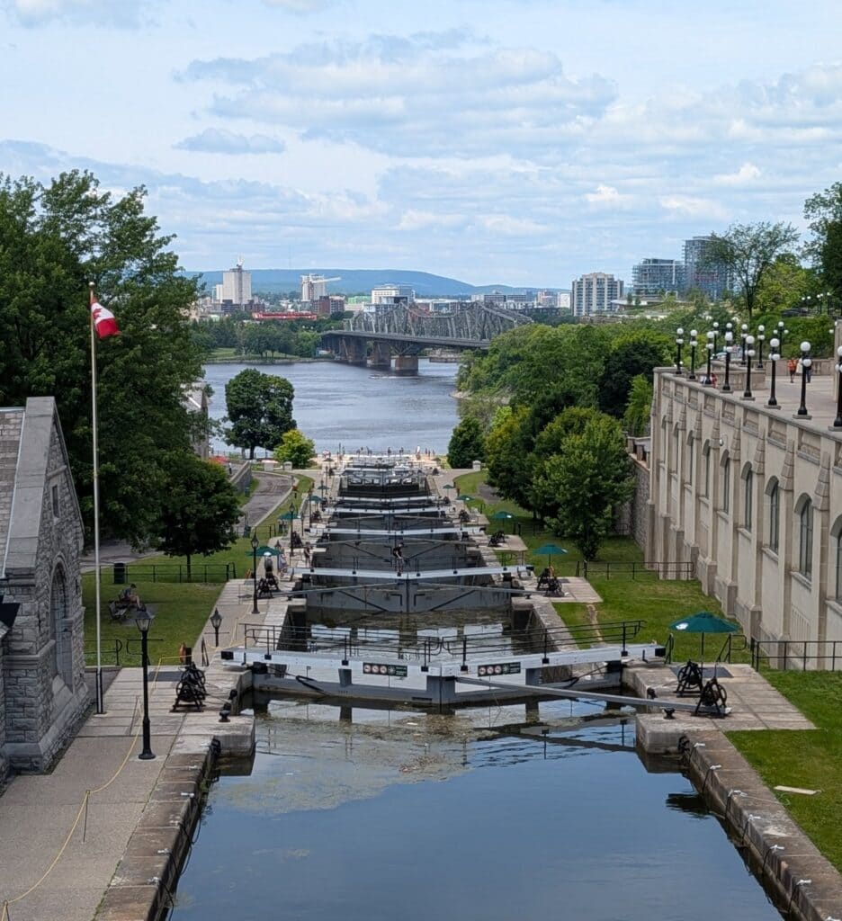 A view of the Rideau Canal locks in downtown Ottawa, with visitors walking along the historic waterway and a bridge spanning the Ottawa River in the background.