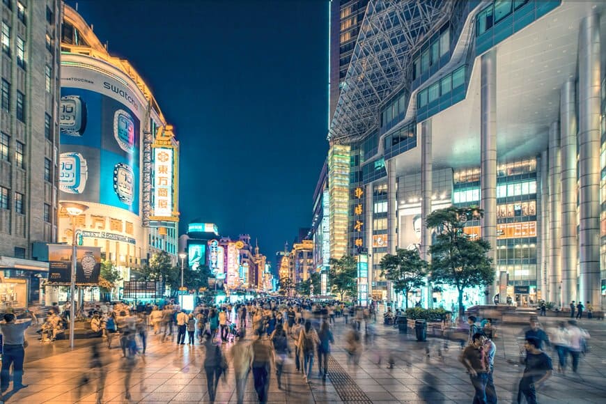 A bustling pedestrian street in Shanghai at night, illuminated by colourful neon signs and shopping malls, with crowds of people walking through the city centre.