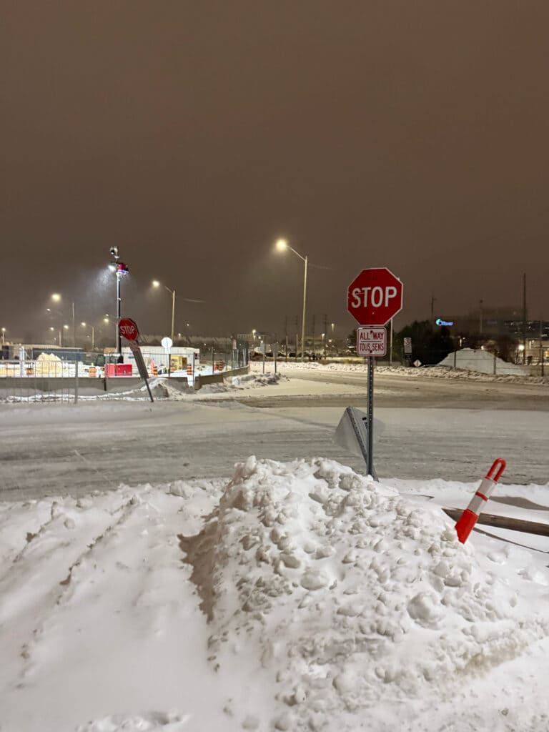 A snow-covered intersection at night, featuring a red stop sign, large snowbanks, and illuminated streetlights under a cloudy winter sky.