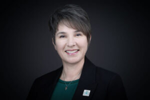 Headshot of a woman wearing a green shirt and black blazer on black background.