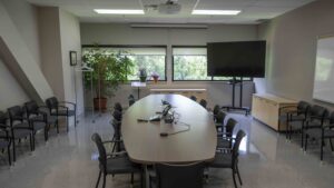 Room with TV, table with 15 chairs, a whiteboard and chairs along the outside wall