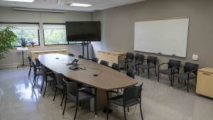 A photo of a big boardroom table with 15 chairs, chairs aligned on the wall, a whiteboard on the wall and TV in the corner. 