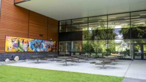 The outdoor space at the Algonquin College Campus. Picnic style tables on a concrete pad, the school's windows in the background, a colourful mural is included in the patio area, a covered area.
