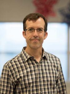 Head shot of a man in a plaid shirt smiling on a beige and white background.
