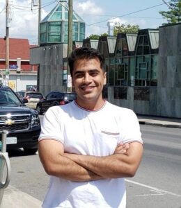 Male with a white shirt and arms crossed smiling with buildings in the background.