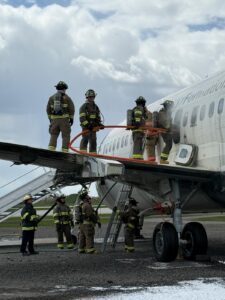 Nine people dressed in firefighter bunker gear taking part in a simulation holding a hose outside of an airplane wing with some people standing on the ground overseeing the scenario.