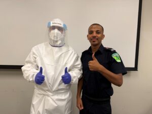 A person dressed in Personal Protective Equipment standing by a male student in a paramedic uniform