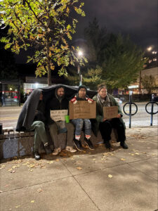Four students sitting on a bench holding signs for the annual 24 Hours of Homelessness Fundraiser for unhoused youth.