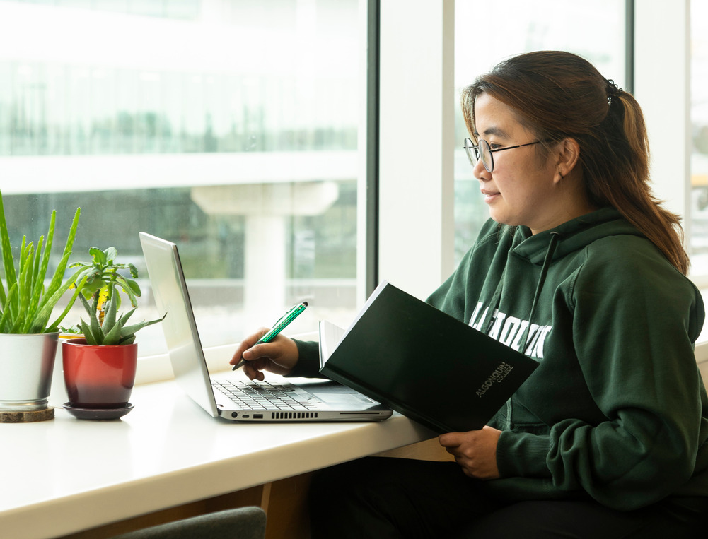 A female student sits at a desk using her laptop and notebook.